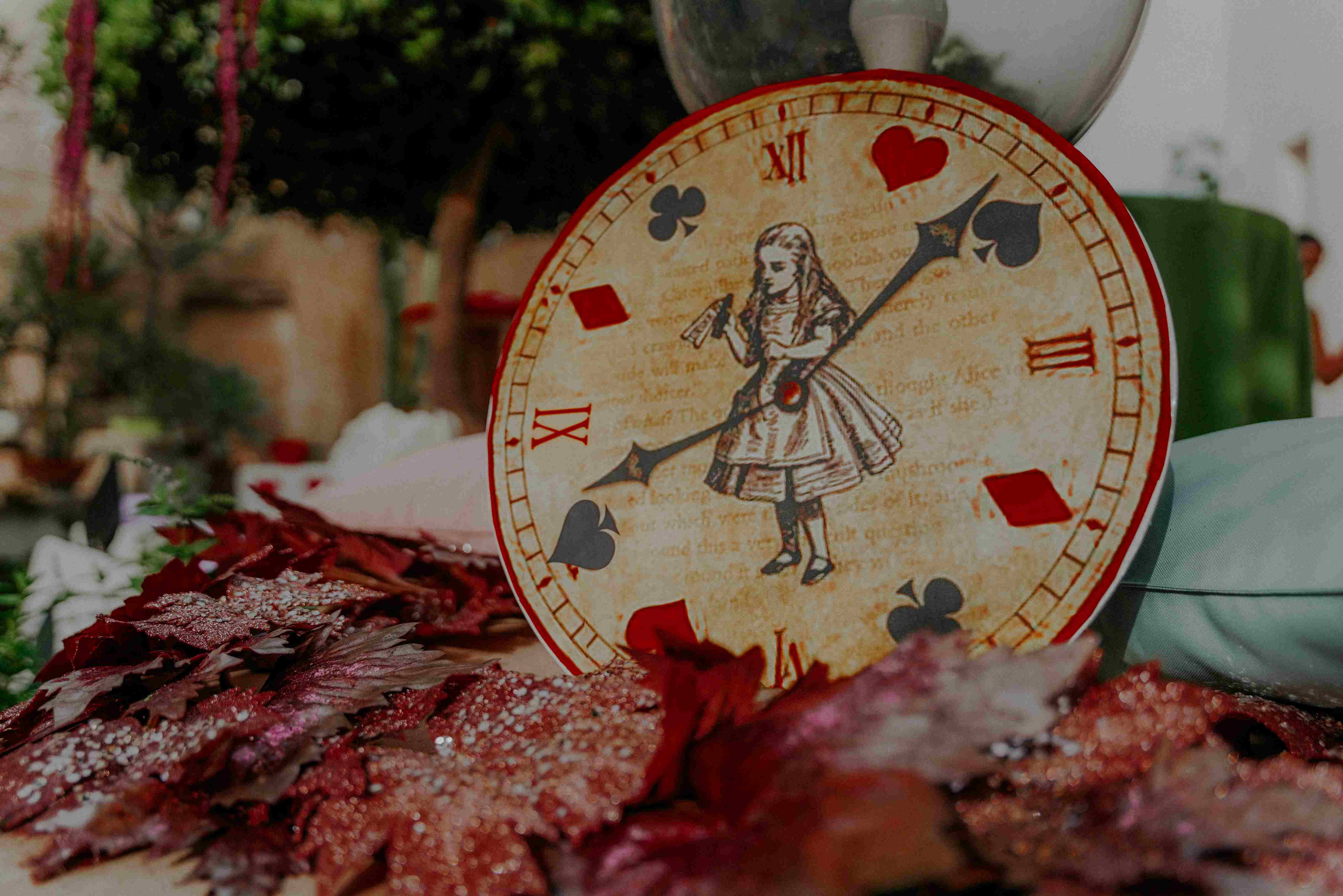 Eine Uhr, auf deren Ziffernblatt Alice im Wunderland zu sehen ist, und die Zeiger zeigen auf Pik, Herz, Karo und Kreuz.
