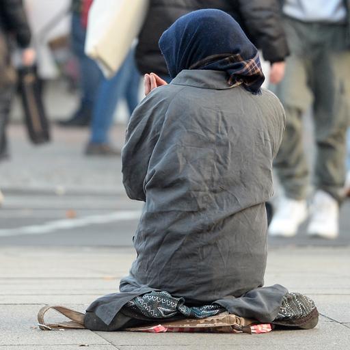 Eine Frau, die man nur von hinten sieht, bettelt inmitten vorbeilaufender Passanten. 