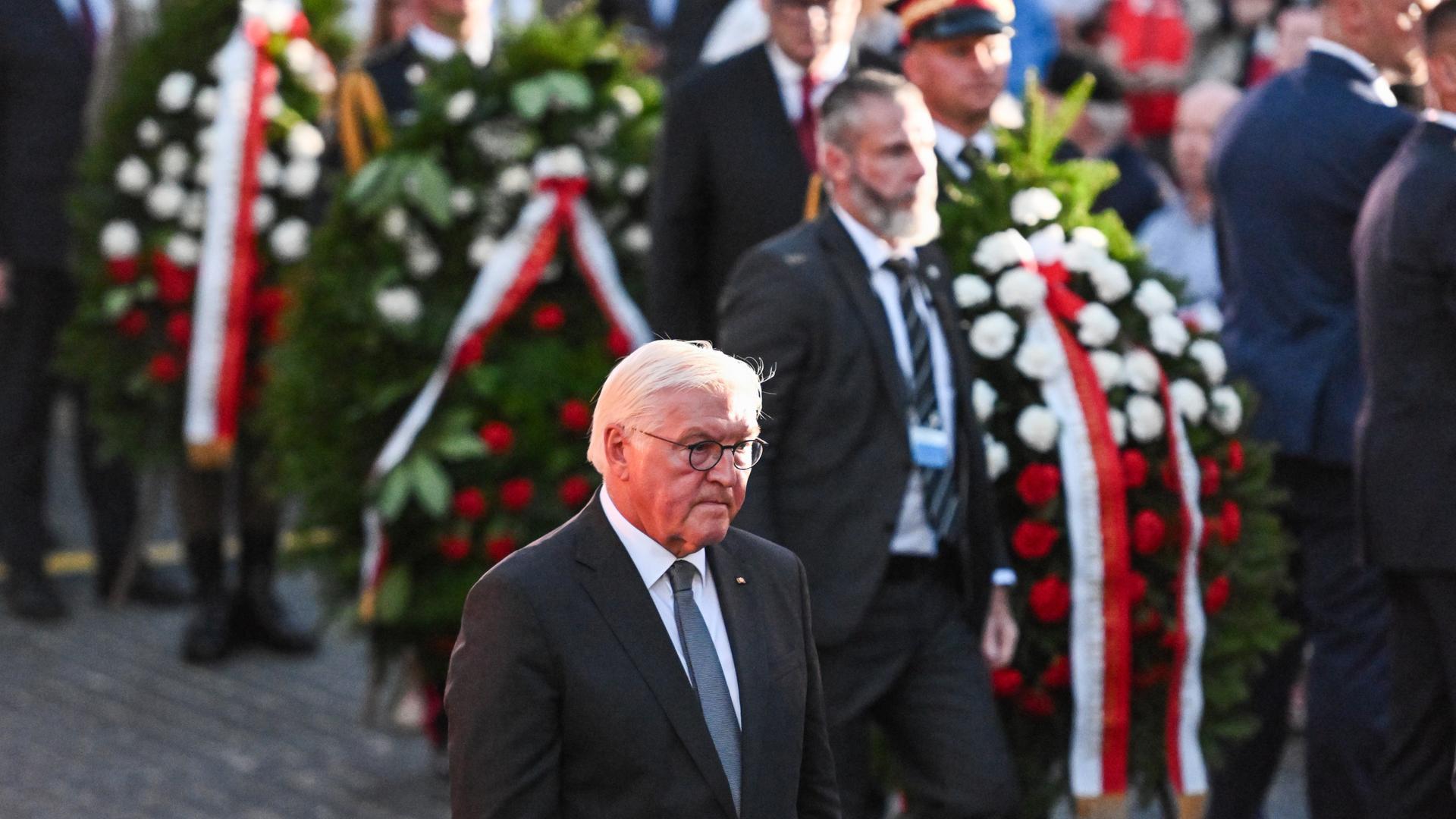 Frank-Walter Steinmeier läuft mit gesenktem ernsten Blick neben weiteren Männern, die Kränze tragen.