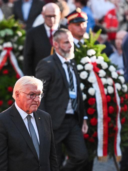 Frank-Walter Steinmeier läuft mit gesenktem ernsten Blick neben weiteren Männern, die Kränze tragen.