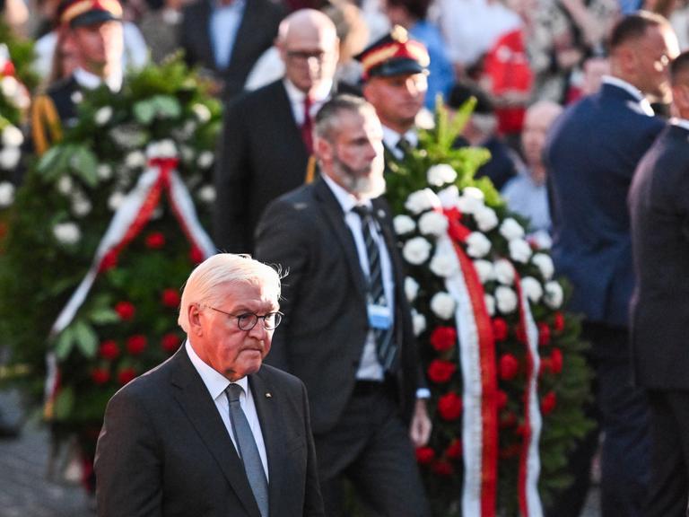 Frank-Walter Steinmeier läuft mit gesenktem ernsten Blick neben weiteren Männern, die Kränze tragen.