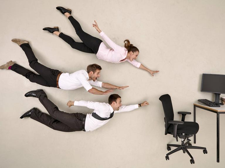 Drei Büromenschen fliegen lachend auf einen Schreibtisch mit Computer zu.