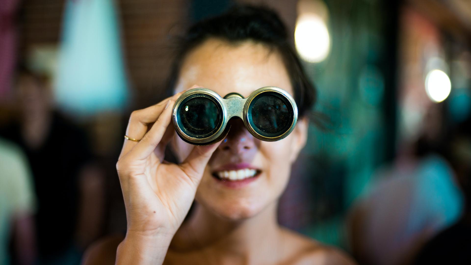 Eine Frau blickt den Betrachtenden direkt durch ein Opernglas an. Sie lächelt. Der Rest des Bildes ist unscharf. Schemenhaft sind Lichter und mindestens zwei weitere Personen zu sehen. 