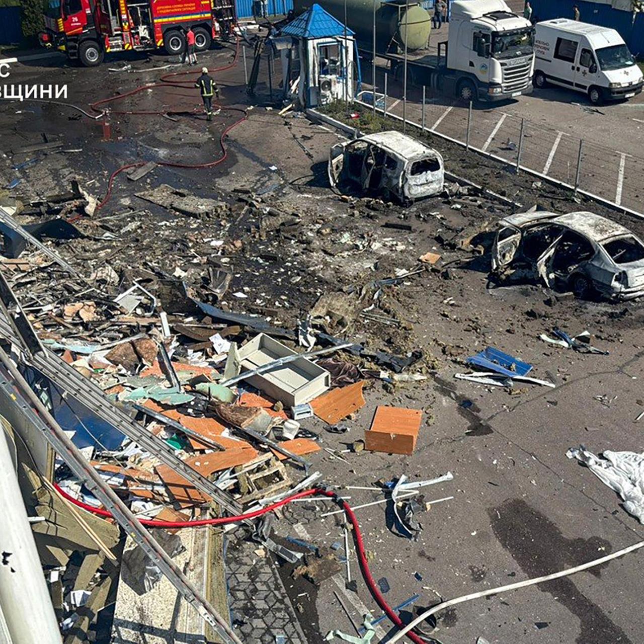 Feuerwehrleute arbeiten auf einem Platz, auf dem Trümmer und zerstörte Autos zu sehen sind.