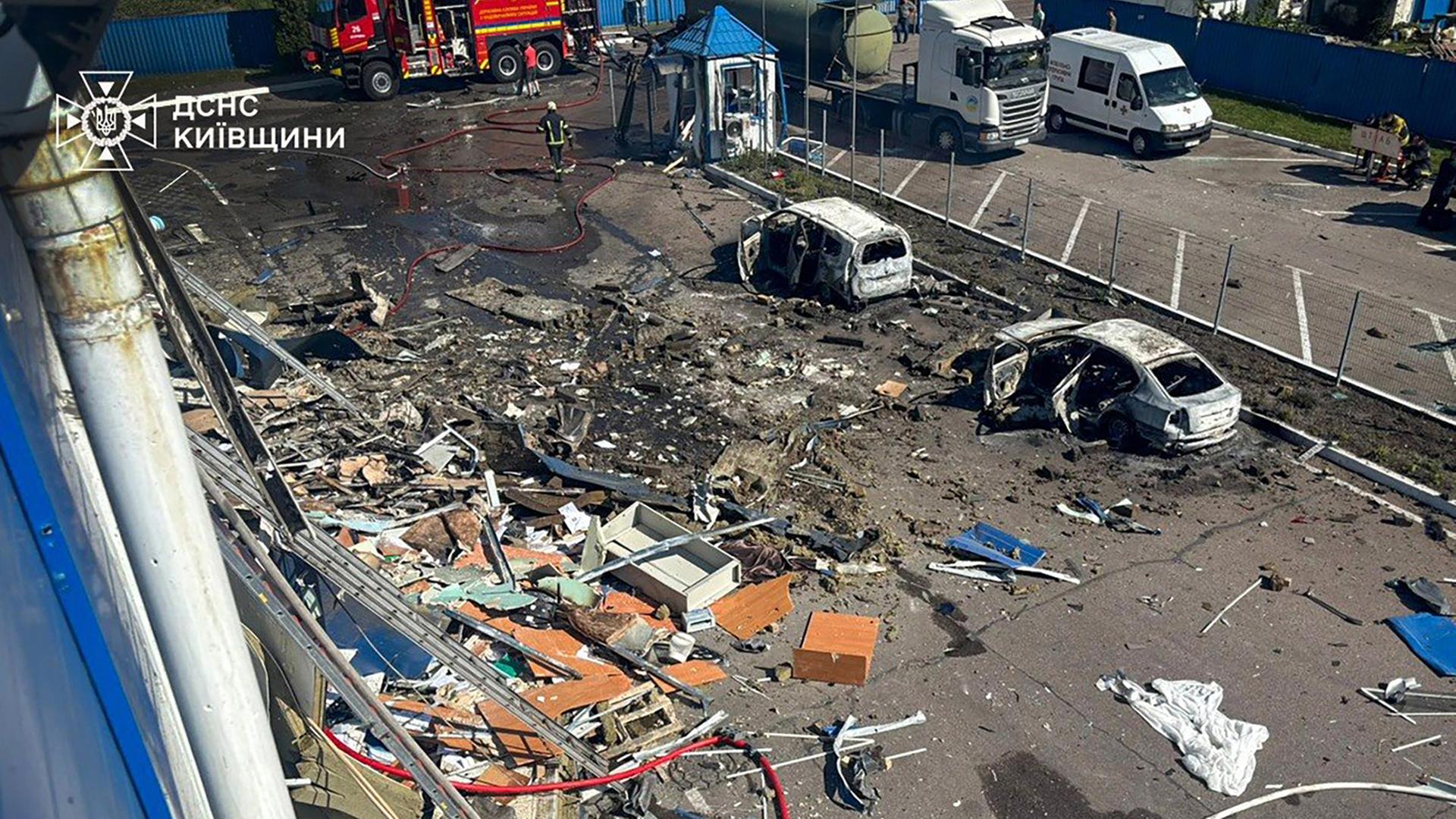 Feuerwehrleute arbeiten auf einem Platz, auf dem Trümmer und zerstörte Autos zu sehen sind.