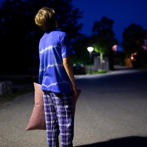 Ein Junge im Schlafanzug steht nachts mit einem Kissen in der Hand auf einer Straße. Während andere tief und fest schlafen, laufen sie oft zur Hochform auf, wenn auch unfreiwillig: Schlafwandler. 