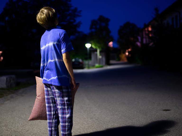 Ein Junge im Schlafanzug steht nachts mit einem Kissen in der Hand auf einer Straße. Während andere tief und fest schlafen, laufen sie oft zur Hochform auf, wenn auch unfreiwillig: Schlafwandler. 