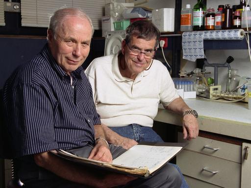 Zwei ältere Männer sitzen nebeneinander in einem Chemielabor und lächeln in die Kamera. Einer von ihnen hält ein aufgeschlagenes Notizbuch auf dem Schoß.