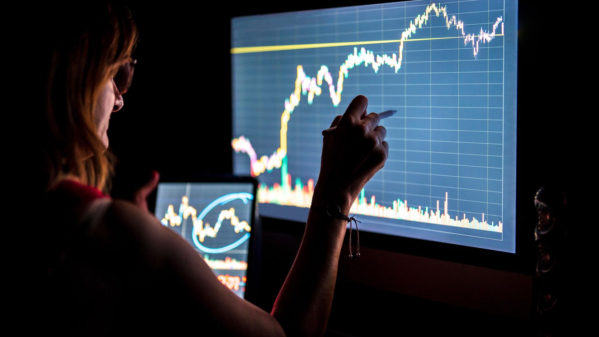 Eine Geschäftsfrau steht in einem dunklen Raum vor einem Monitor und hält ein Tablett in der Hand. Auf beiden Bildschirmen sieht man Finanzkurven