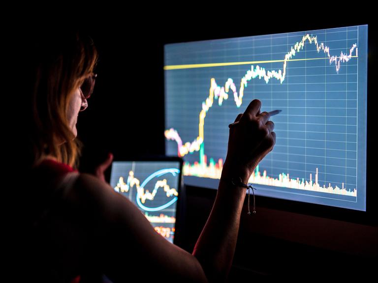 Eine Geschäftsfrau steht in einem dunklen Raum vor einem Monitor und hält ein Tablett in der Hand. Auf beiden Bildschirmen sieht man Finanzkurven