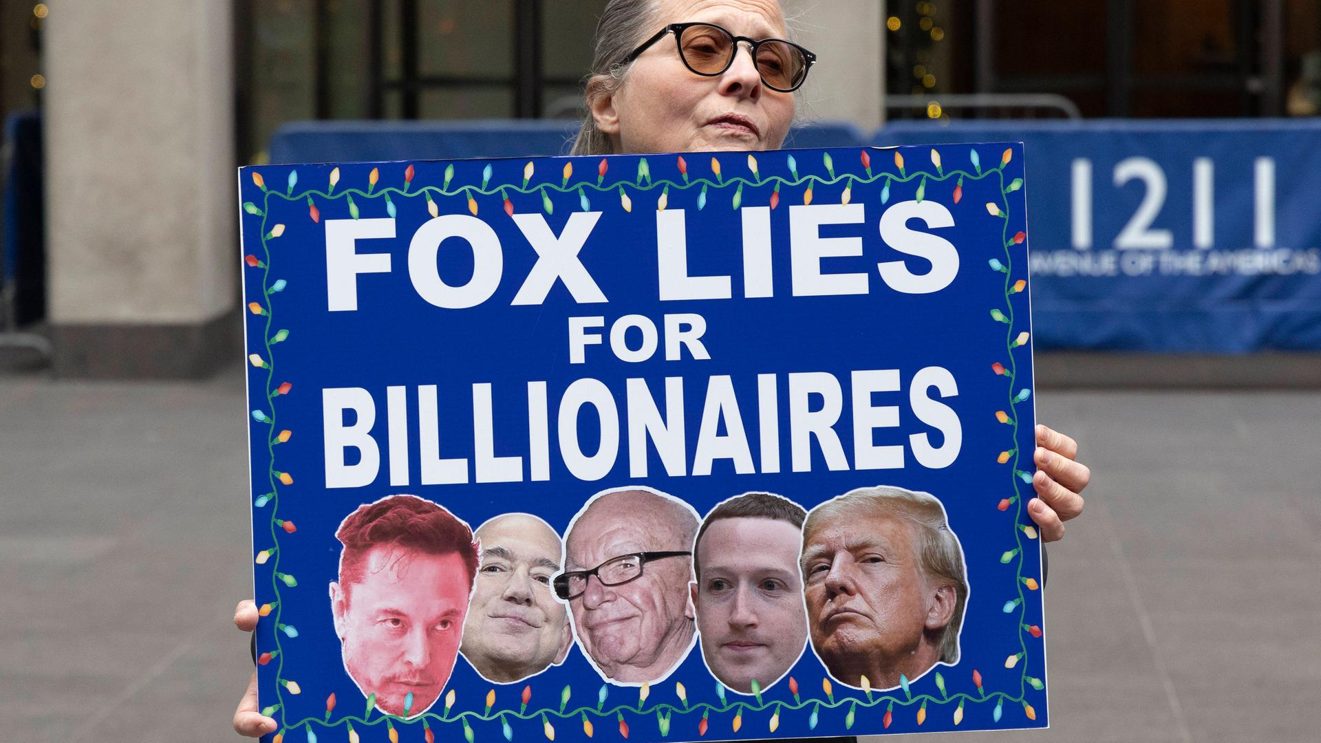 Eine Demonstrantin hält ein Schild mit der Aufschrift „FOX LIES FOR BILLIONAIRES” (FOX LÜGT FÜR MILLIARDÄRE) mit Bildern von Musk, Bezos, Murdoch, Zuckerberg und Trump vor dem Fox News Channel in New York City, USA, November 2025.