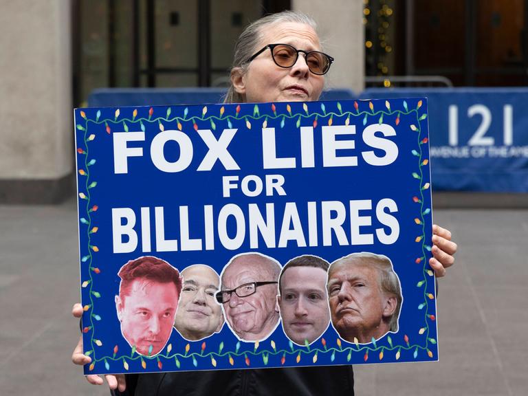 Eine Demonstrantin hält ein Schild mit der Aufschrift „FOX LIES FOR BILLIONAIRES” (FOX LÜGT FÜR MILLIARDÄRE) mit Bildern von Musk, Bezos, Murdoch, Zuckerberg und Trump vor dem Fox News Channel in New York City, USA, November 2025.