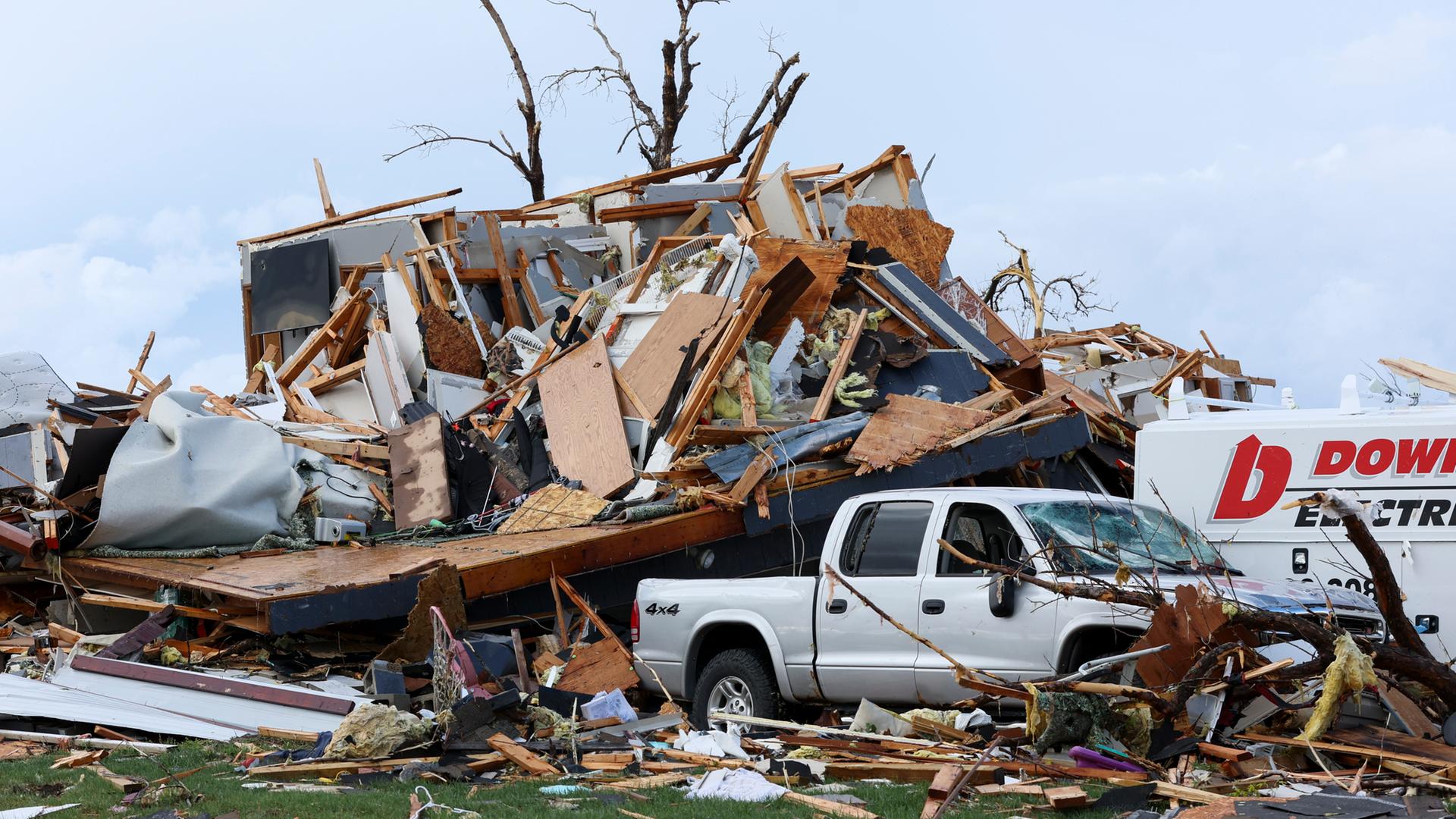 Ein Haus liegt nach einem Tornado in Trümmern. Hinter den Trümmern sieht man die kahlen Äste eines Baumes. Im Vordergrund ist ein Auto, das teilweise mit Brettern und Ästen bedeckt ist.