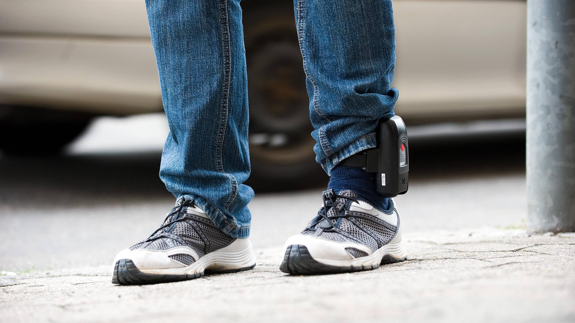 Detailaufnahme der Beine eines Mannes in Jeans und Turnschuhen, der eine elektronische Fußfessel trägt.