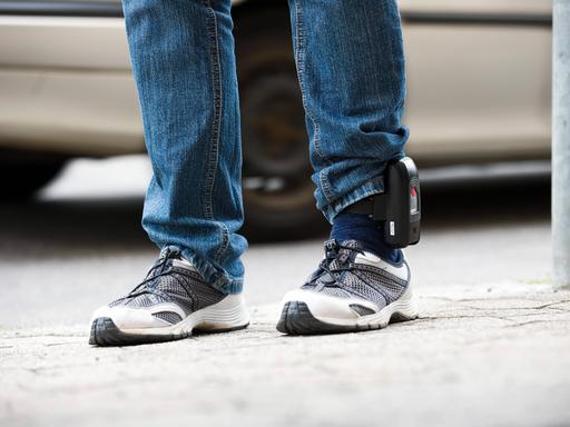 Detailaufnahme der Beine eines Mannes in Jeans und Turnschuhen, der eine elektronische Fußfessel trägt.