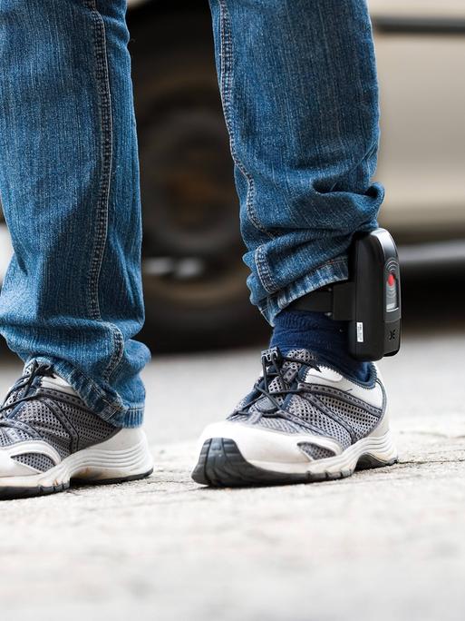 Detailaufnahme der Beine eines Mannes in Jeans und Turnschuhen, der eine elektronische Fußfessel trägt.