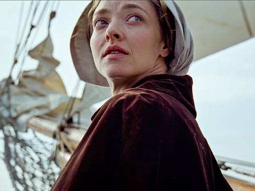 Die Hauptdarstellerin Amanda Seyfried in ihrer Rolle als Ann Lee dreht sich um. Im Hintergrund ist der Mast eines Bootes zu sehen, sie trägt eine traditionelle Kopfbedeckung. 