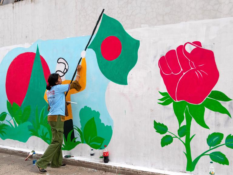 Graffiti auf einer Wand in Bangladesch: Eine Studentin malt einen Demonstranten, der eine Flagge von Bangladesch hält, aus einer Rose wächst eine Faust. Graffiti auf einer Wand in Bangladesch: Eine Studentin malt einen Demonstranten, der eine Flagge von Bangladesch hält, aus einer Rose wächst eine Faust.