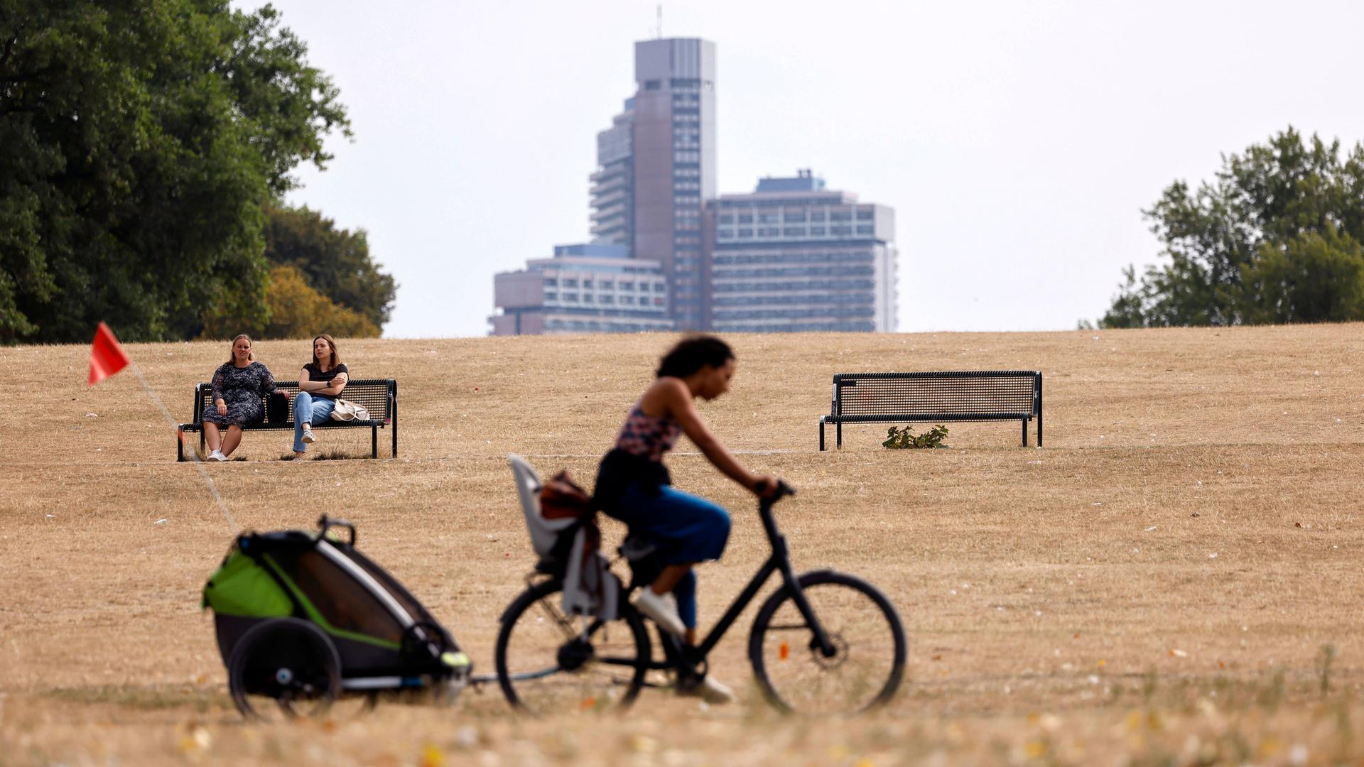 Eine Frau fährt mit einem Fahrrad mit Kindersitz auf dem Gepäckträger und Fahrradanhänger durch einen Park, dessen Rasenfläche braun verbrannt ist. Links sitzen im Hintergrund zwei Frauen auf einer Parkbank, noch weiter hinten ist ein Hochhaus zu sehen.