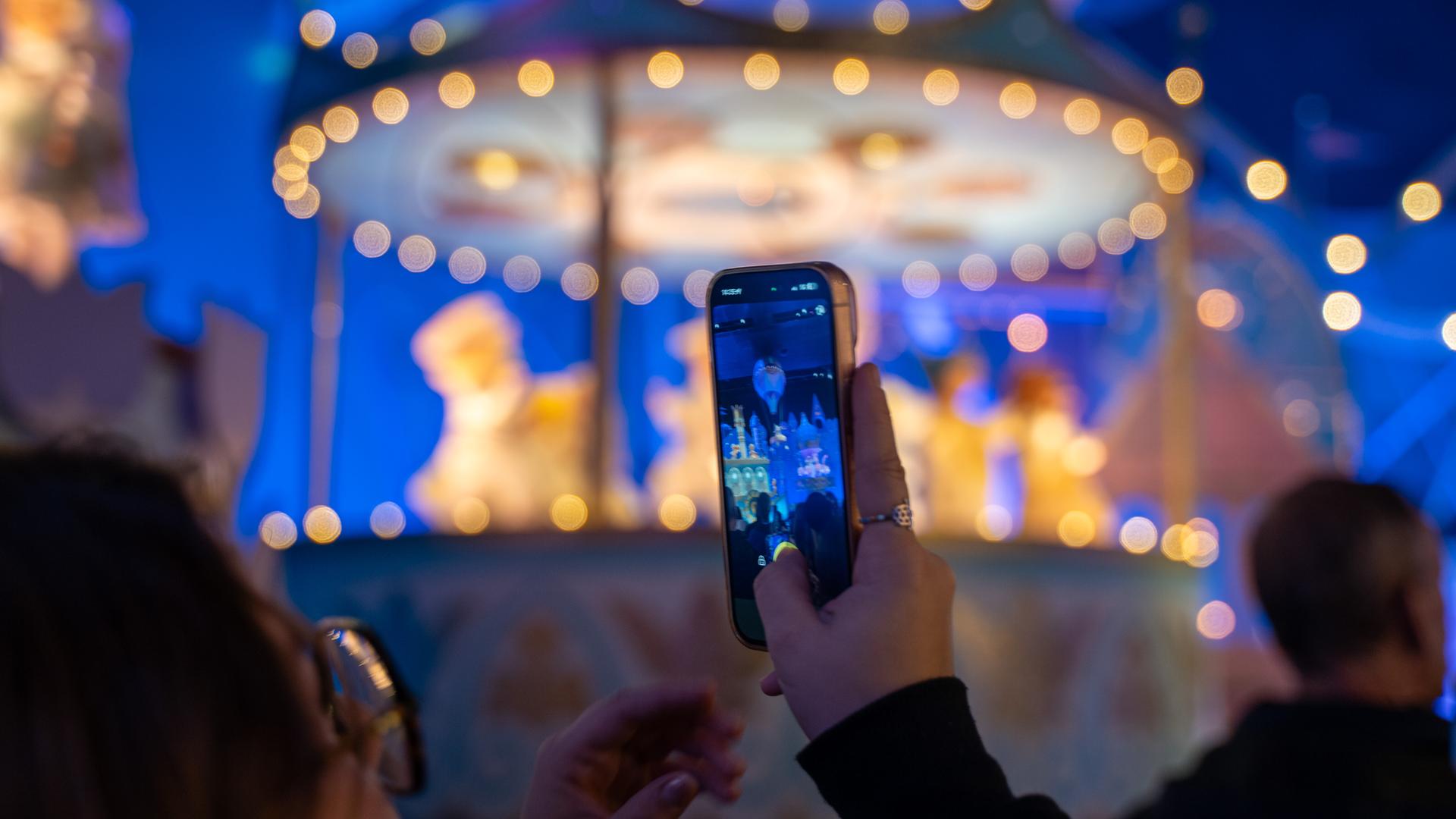 In einen Freizeitpark hält ein Kind ein Smartphone in die Höhe und macht ein Foto. Im Hintergrund ist verschwommen ein Fahrgeschäft.