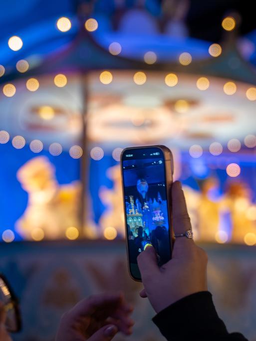In einen Freizeitpark hält ein Kind ein Smartphone in die Höhe und macht ein Foto. Im Hintergrund ist verschwommen ein Fahrgeschäft.