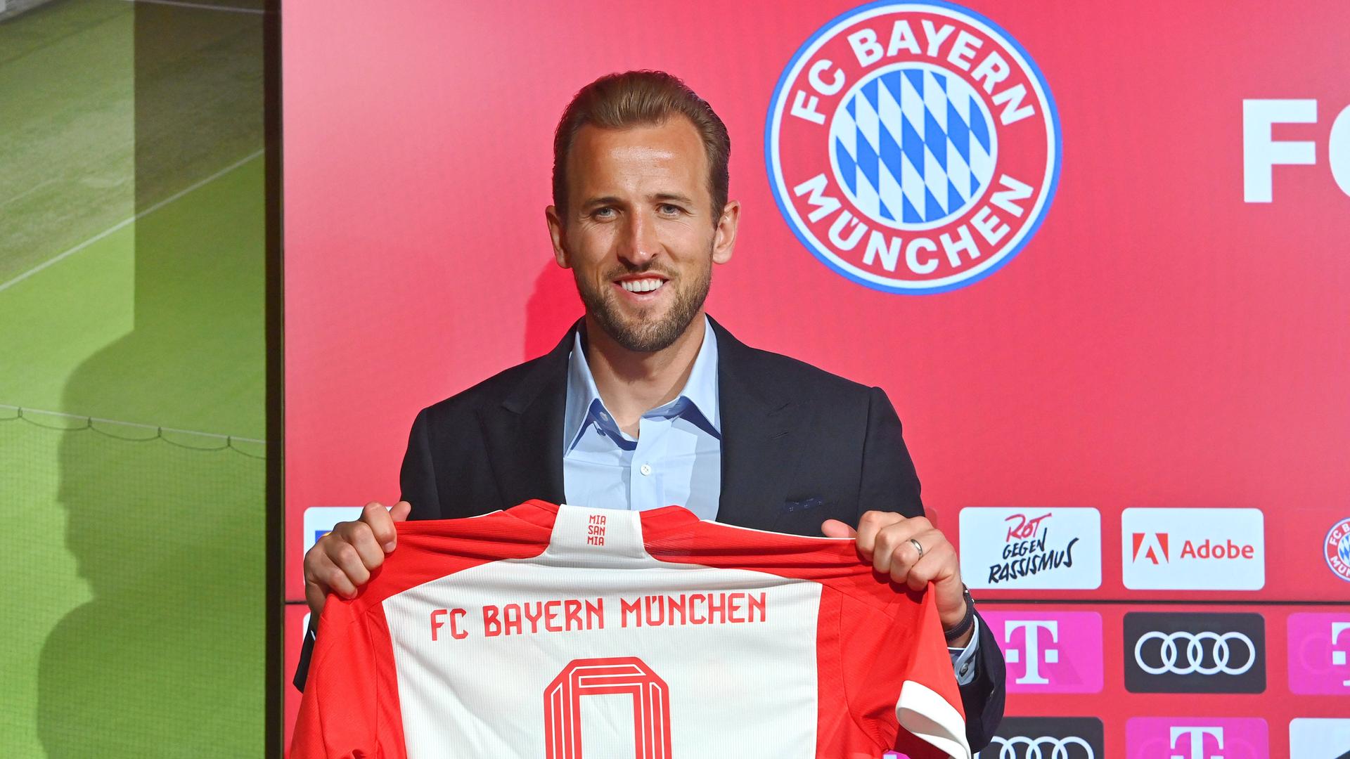 Fußballspieler Harry Kane im Anzug hält sein Trikot vom FC Bayern München in den Händen.