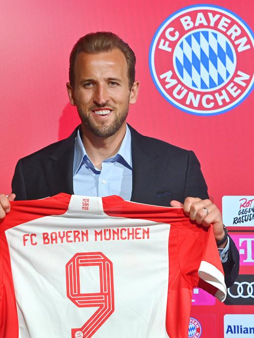 Fußballspieler Harry Kane im Anzug hält sein Trikot vom FC Bayern München in den Händen.