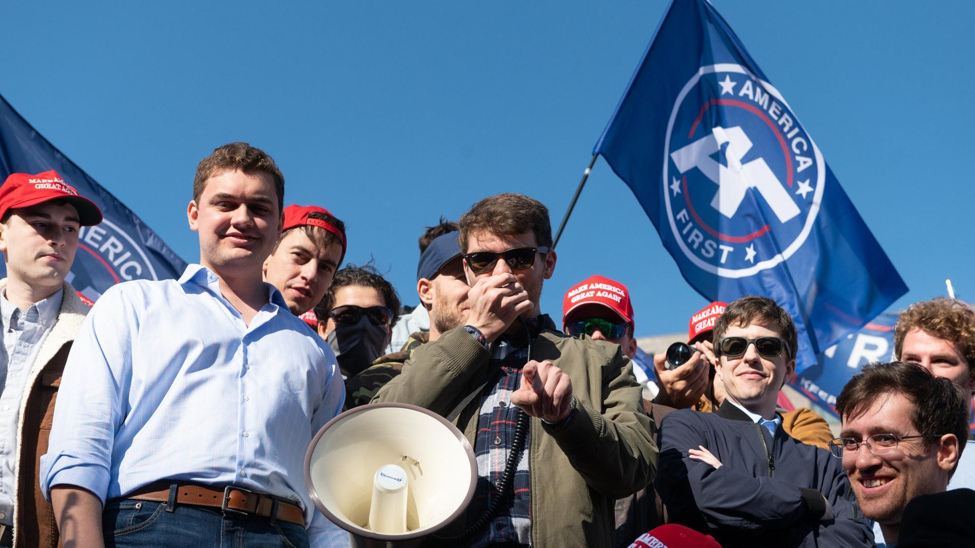 Nick Fuentes, Führer der Groypers, mit seinen Anhängern. Ein Sprecher hält ein Megafon in der Hand, um ihn herum stehen junge Männer und grinsen. 