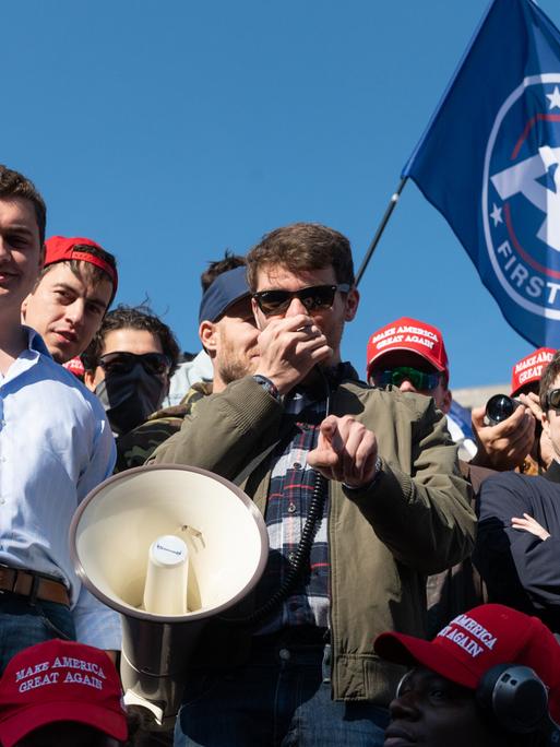 Nick Fuentes, Führer der Groypers, mit seinen Anhängern. Ein Sprecher hält ein Megafon in der Hand, um ihn herum stehen junge Männer und grinsen. 