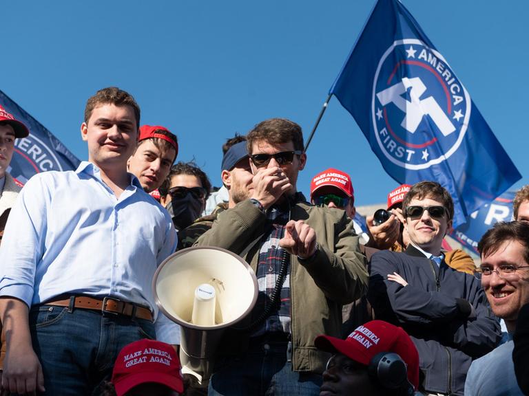 Nick Fuentes, Führer der Groypers, mit seinen Anhängern. Ein Sprecher hält ein Megafon in der Hand, um ihn herum stehen junge Männer und grinsen. 