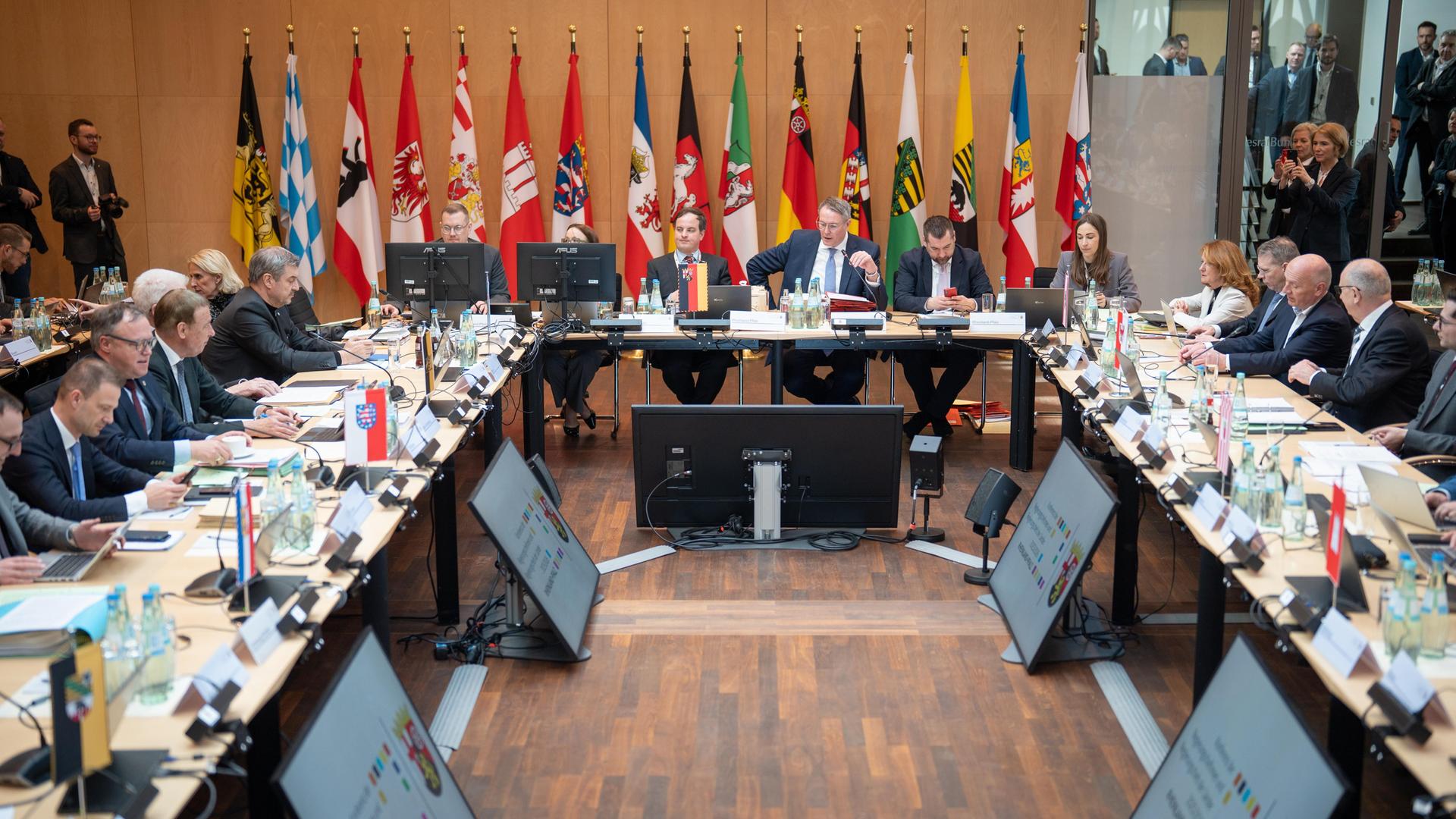 Die Ministerpräsidenten und ihre Mitarbeiter sitzen an hufeisenfömig angeordneten Tischen in einem Raum. Hinten an der Wand stehen die Fahnen der Bundesländer. 