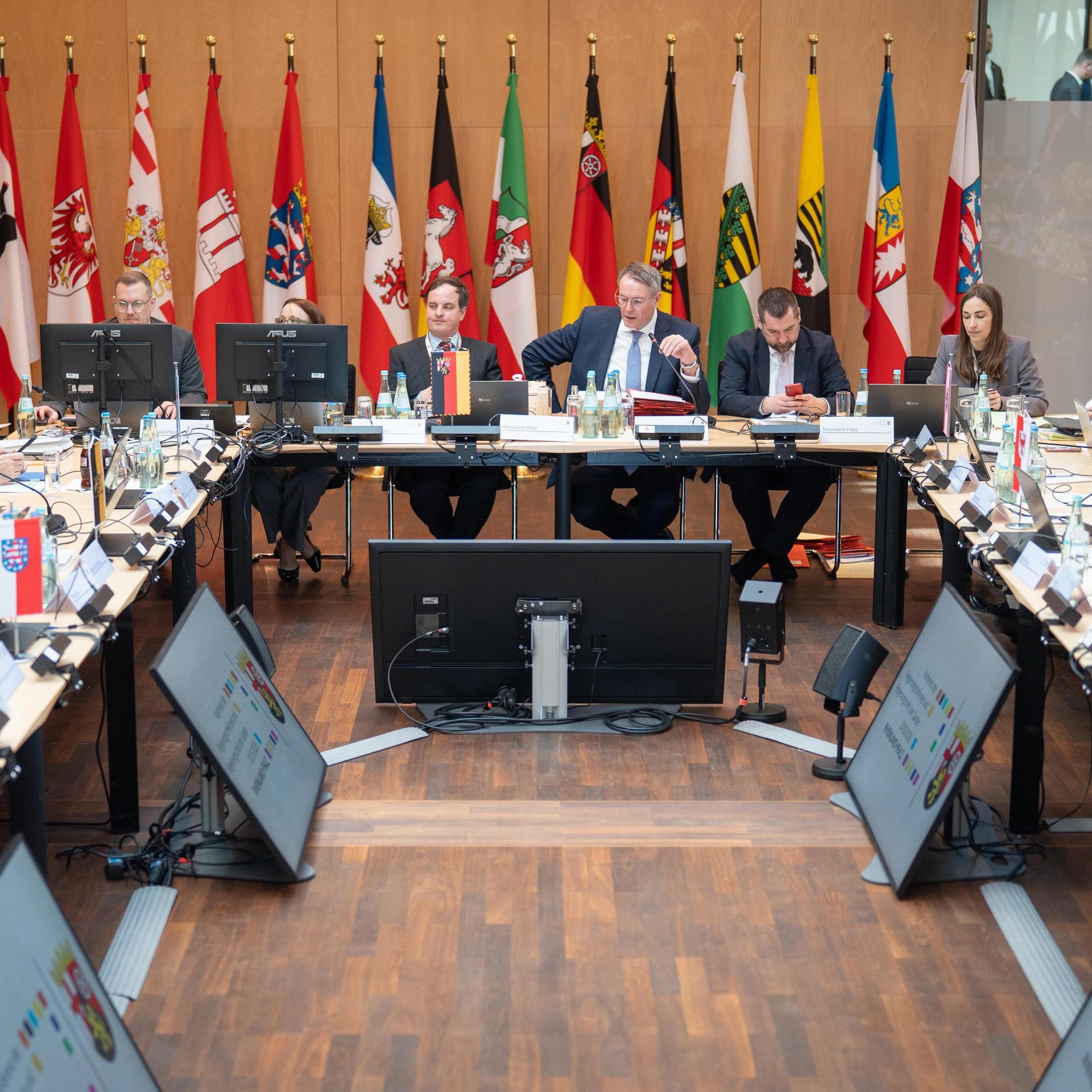 Die Ministerpräsidenten und ihre Mitarbeiter sitzen an hufeisenfömig angeordneten Tischen in einem Raum. Hinten an der Wand stehen die Fahnen der Bundesländer. 
