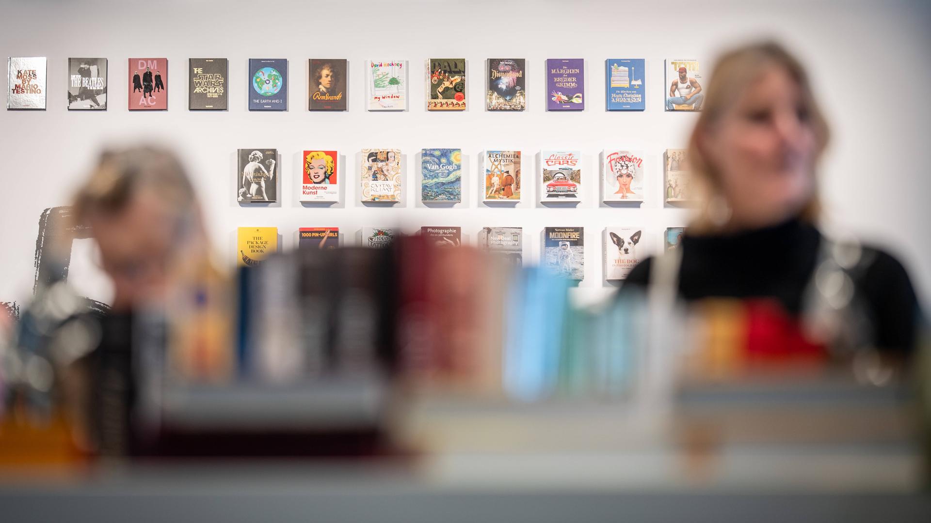 Eine weiße Wand mit vielen Büchern ist im Fokus, davor stehen zwei Personen im Vordergrund verschwommen Eine weiße Wand mit vielen Büchern ist im Fokus, davor stehen zwei Personen im Vordergrund verschwommen