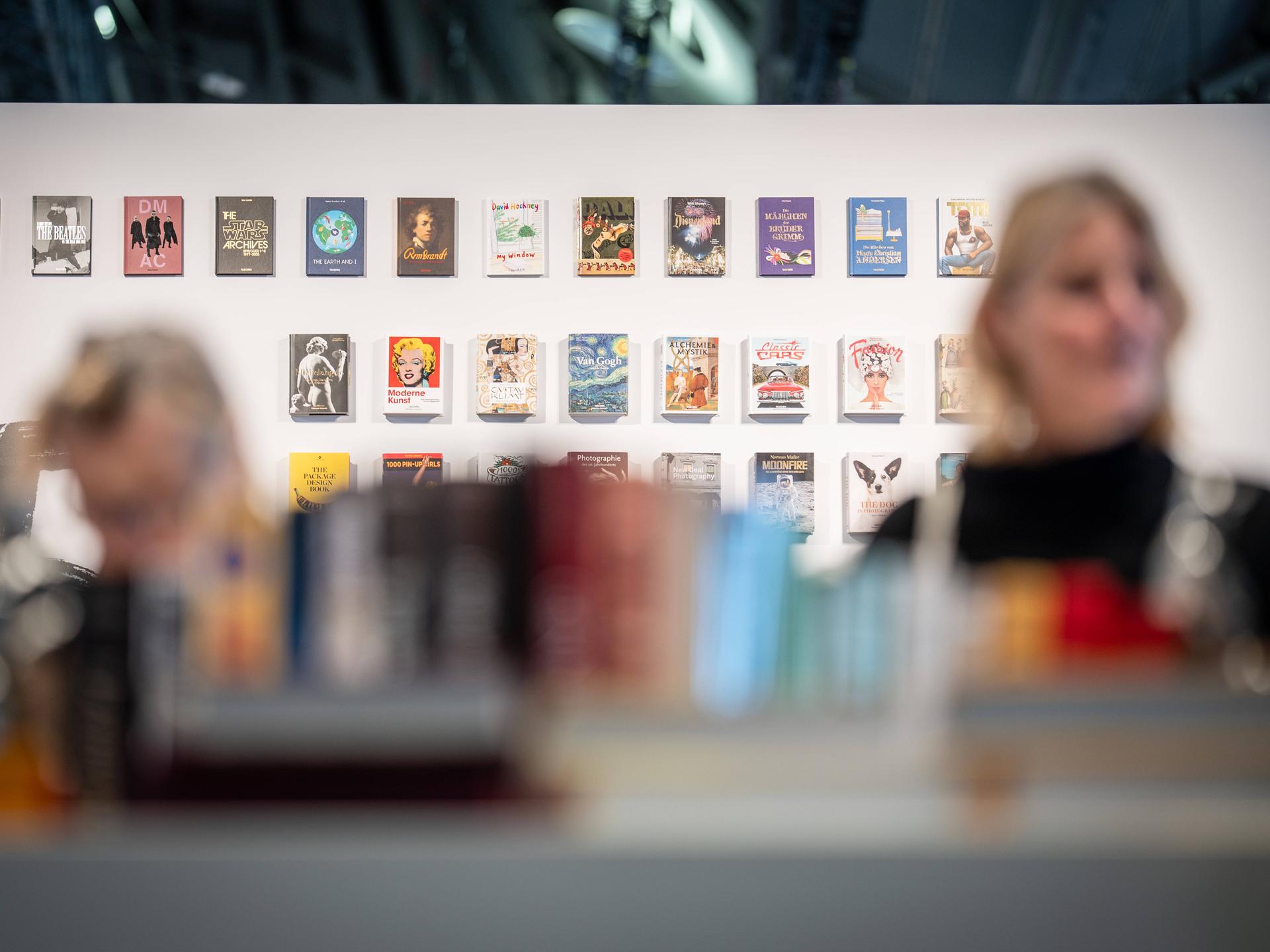 Eine weiße Wand mit vielen Büchern ist im Fokus, davor stehen zwei Personen im Vordergrund verschwommen