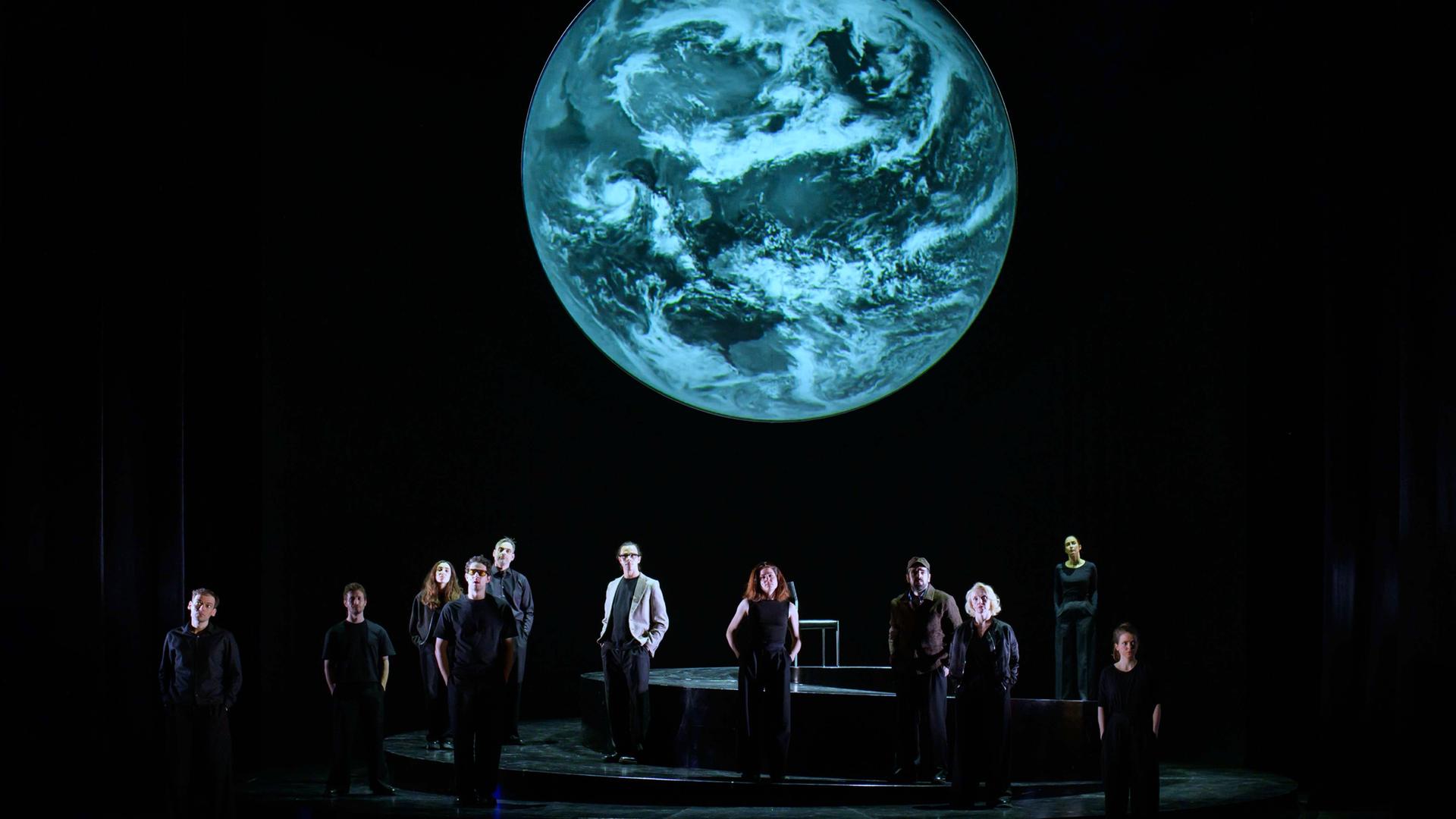 Szenenfoto aus der Produktion "Wir sind das Volk" am Deutschen Nationaltheater Weimar: Schauspielende auf einer dunklen Bühne blicken Richtung Publikum, hinter ihnen eine Projektion der Erdkugel. Szenenfoto aus der Produktion "Wir sind das Volk" am Deutschen Nationaltheater Weimar: Schauspielende auf einer dunklen Bühne blicken Richtung Publikum, hinter ihnen eine Projektion der Erdkugel.