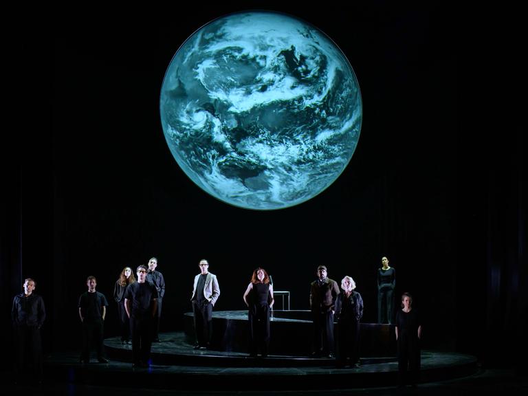 Szenenfoto aus der Produktion "Wir sind das Volk" am Deutschen Nationaltheater Weimar: Schauspielende auf einer dunklen Bühne blicken Richtung Publikum, hinter ihnen eine Projektion der Erdkugel. Szenenfoto aus der Produktion "Wir sind das Volk" am Deutschen Nationaltheater Weimar: Schauspielende auf einer dunklen Bühne blicken Richtung Publikum, hinter ihnen eine Projektion der Erdkugel.