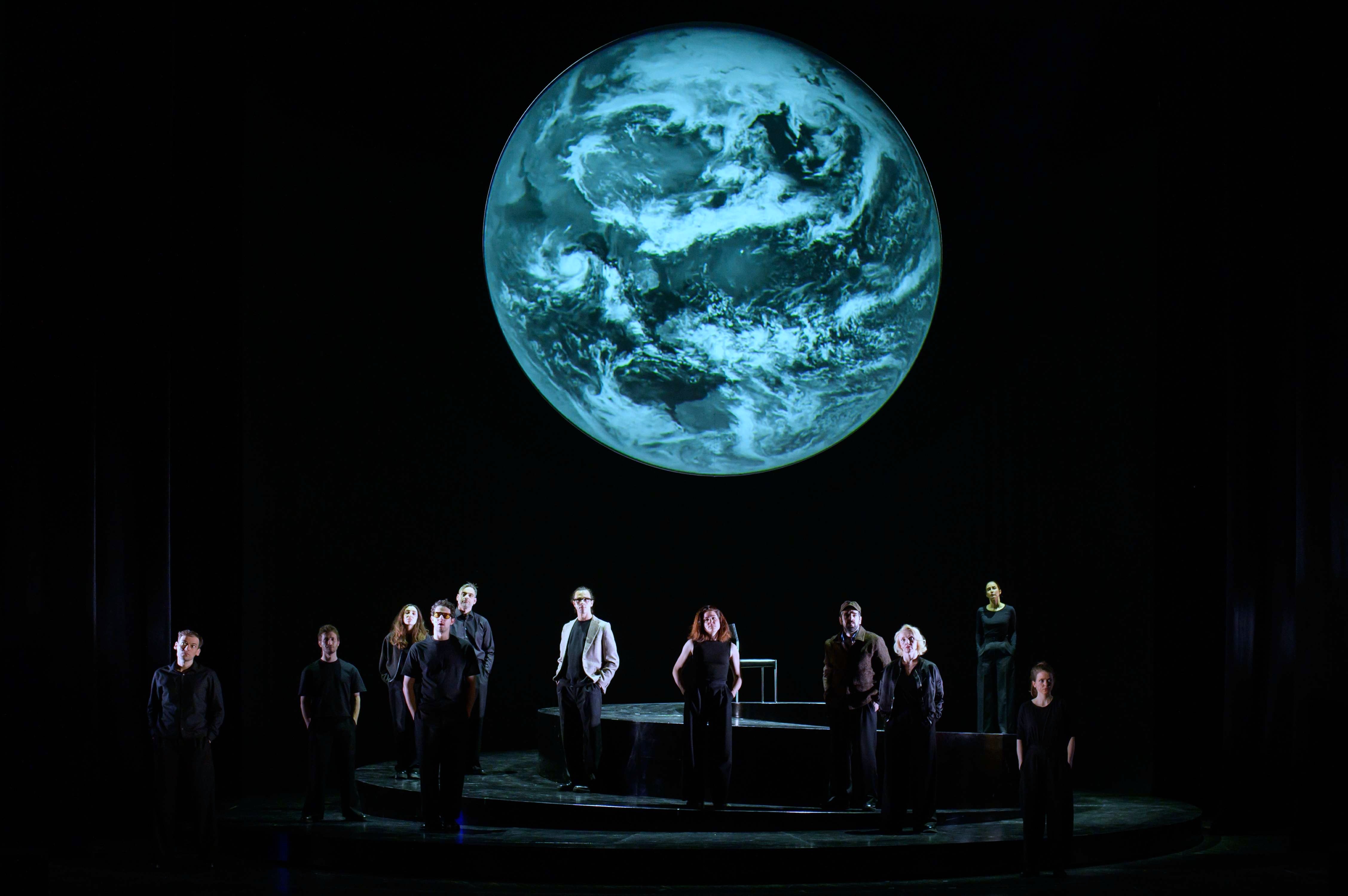 Szenenfoto aus der Produktion "Wir sind das Volk" am Deutschen Nationaltheater Weimar: Schauspielende auf einer dunklen Bühne blicken Richtung Publikum, hinter ihnen eine Projektion der Erdkugel.