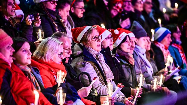 Mehrere Menschen sitzen im Stadion. Einige Tragen Weihnachts-Mützen. Andere halten eine Kerze. Sie singen Weihnachts-Lieder. 