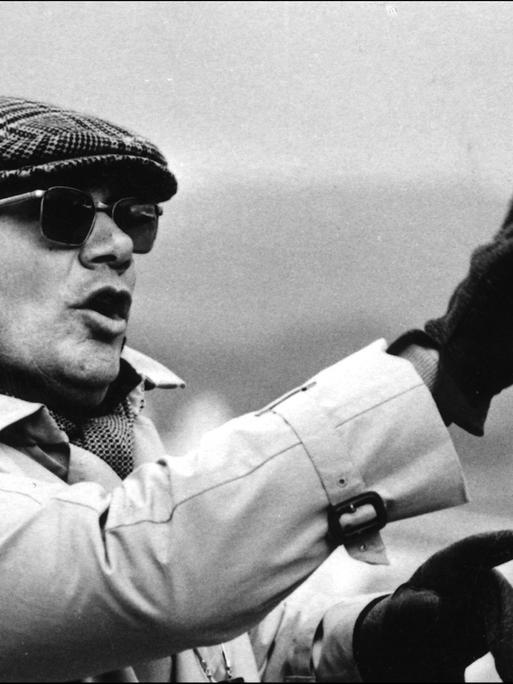 Der italienische Filmregisseur und Drehbuchautor Francesco Rosi gestikuliert und trägt eine Mütze, Sonnenbrille, Handschuhe und einen Regenmantel in den 1970er-Jahren