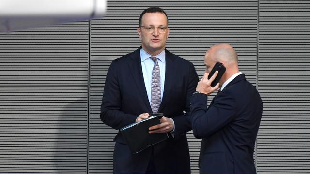 Jens Spahn steht bei der 19. Sitzung des 21. Deutschen Bundestages im Reichstagsgebäude und hält einen Ordner, daneben ein telefonierender Mann