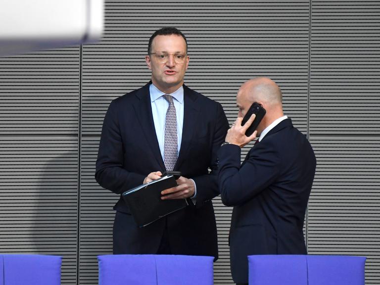Jens Spahn steht bei der 19. Sitzung des 21. Deutschen Bundestages im Reichstagsgebäude und hält einen Ordner, daneben ein telefonierender Mann