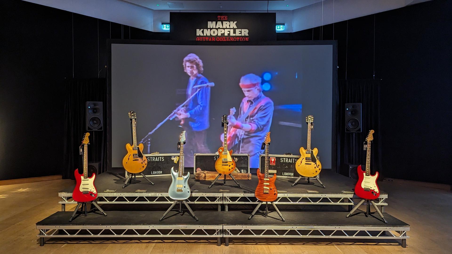 Mark Knopflers Gitarren werden im Auktionshaus Christie's auf einer Bühne ausgestellt, dahinter ein Bild mit Musiker bei einem Konzert