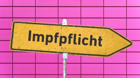 Ein gelbes, zerkratztes Hinweisschild mit der Aufschrift "Impfpflicht" vor einer rosa Kachelwand. 