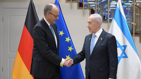 Friedrich Merz und Benjamin Nethanjahu stehen vor der deutschen und der israelischen Flagge und geben sich die Hand.