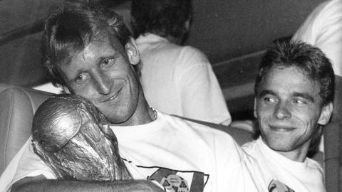 Andreas Brehme hält vor der Abfahrt des DFB-Mannschaftsbusses in Rom den eroberten WM-Pokal im Arm, Teamgefährte Thomas Hässler schaut lächelnd zu. 