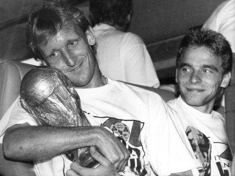 Andreas Brehme hält vor der Abfahrt des DFB-Mannschaftsbusses in Rom den eroberten WM-Pokal im Arm, Teamgefährte Thomas Hässler schaut lächelnd zu. 