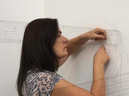 Christiane Löhr steht an einer Wand, in der im Kreis Nadeln stecken, von diesen Nadeln ziehen sich parallele Fäden in den Raum. Sie arbeitet konzentriert und bewegt Nadel und Faden durch die aufgespannten Fäden.