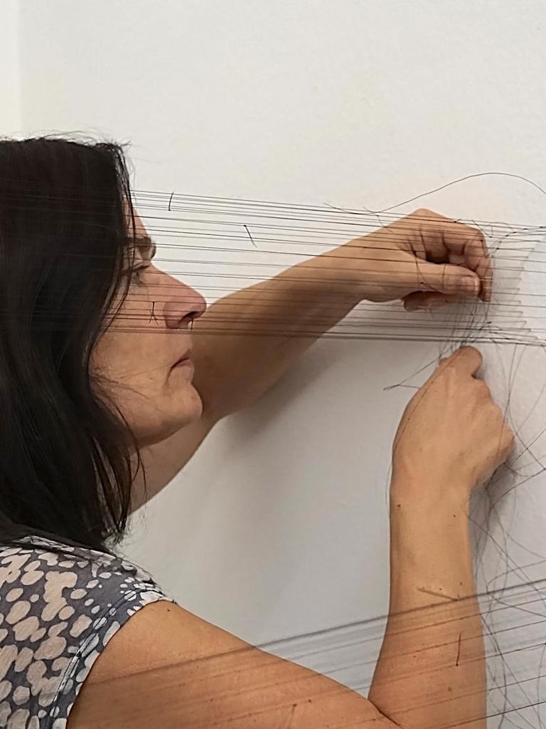 Christiane Löhr steht an einer Wand, in der im Kreis Nadeln stecken, von diesen Nadeln ziehen sich parallele Fäden in den Raum. Sie arbeitet konzentriert und bewegt Nadel und Faden durch die aufgespannten Fäden.