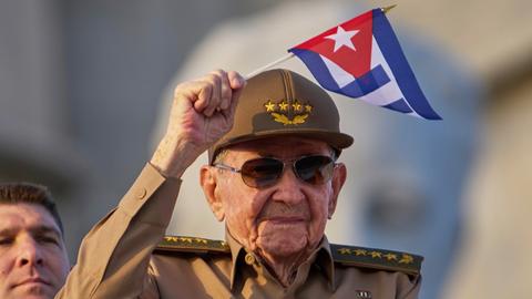 Castro trägt eine Uniform und Sonnenbrille. Er schwenkt eine kleine kubanische Flagge 