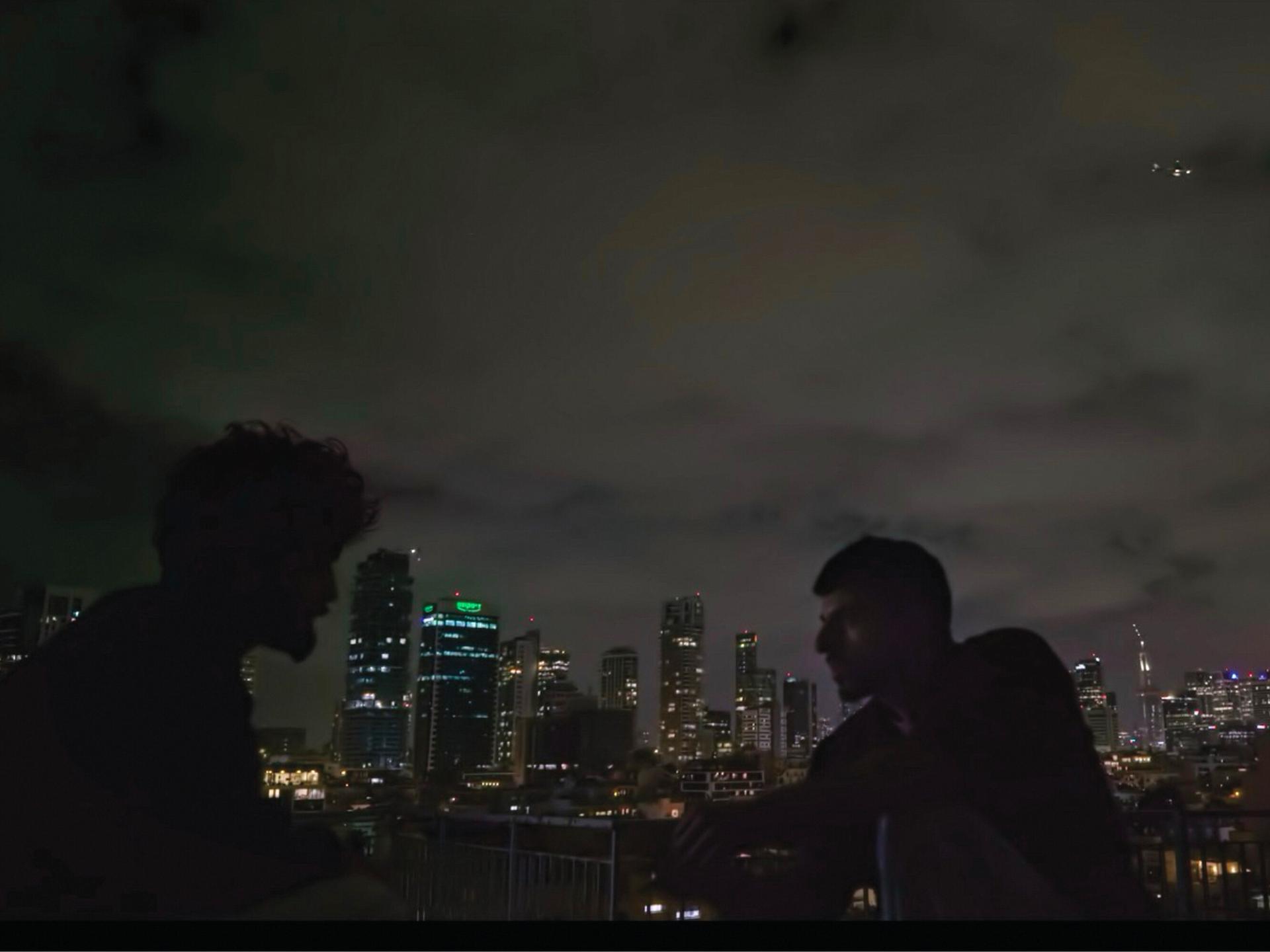 Zwei Männer sind im Profil zu erkennen. Sie sitzen im Dunkeln draußen. Im Hintergrund ist die Skyline von Tel Aviv zu erkennen. Es handelt sich um eine Szene aus der Doku "NAZA". Zwei Männer sind im Profil zu erkennen. Sie sitzen im Dunkeln draußen. Im Hintergrund ist die Skyline von Tel Aviv zu erkennen. Es handelt sich um eine Szene aus der Doku "NAZA".